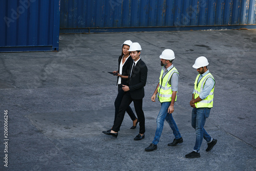Three professionals conduct tablet-based inspection of shipping containers within an industrial warehouse environment while wearing hard hats and safety vests for enhanced workplace protection.