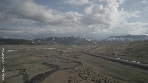 Low altitude drone flight along mountain river and dirt road in scenic spring valley against snowy peaks. Travel journey. RAW flat profile uncorrected video footage.