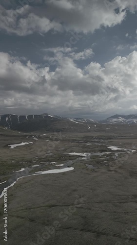 Aerial pull out shot of winding mountain river flowing through spring steppe valley against snowy peaks. Scenic landscape panorama. RAW flat profile uncorrected video.