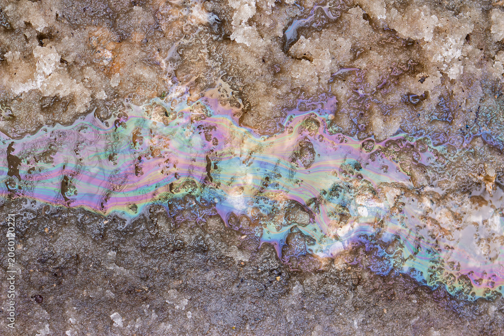 Naklejka premium Iridescent oil stain creating a colorful rainbow sheen on melting dirty spring snow, showing environmental pollution and chemical waste runoff on the city road surface during seasonal thaw