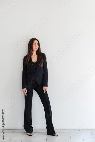 Schilderij op canvas Woman looking side in black formal pantsuit or suit stands against white wall in studio