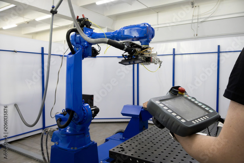 Operator uses handheld teach pendant to program or control a blue industrial robot arm. The manipulator is equipped with complex tooling and suspended above a perforated work surface. Clean white wall