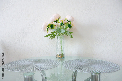 Elegant bouquet of pale pink peonies sits in a clear, textured glass vase atop a sleek glass table. Two silver patterned placemats frame the centerpiece. Soft lighting and clean white wall create a tr