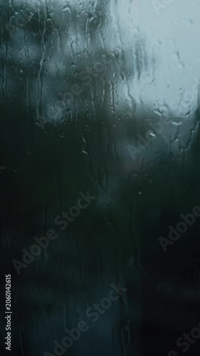 Raindrops stream down a window pane during a heavy downpour, creating a blurry and distorted view of the green trees outside. This moody shot captures the cozy, melancholy atmosphere of a stormy day.