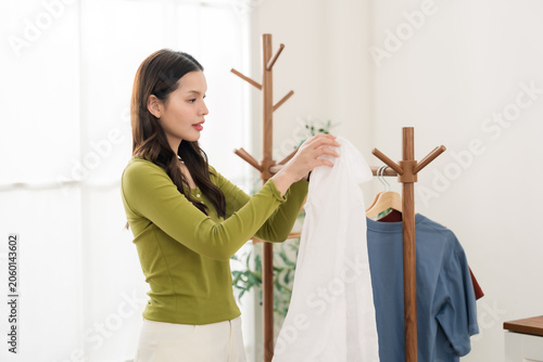 Young asian woman hangs white shirt on wooden clothes rack near bright window, weekend laundry drying routine at home, cozy apartment lifestyle with tidy domestic care and calm homemaking task