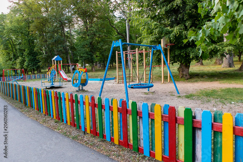 Children's playground surrounded by a colorful wooden fence, featuring swings, slide, climbing structures, and green park scenery.