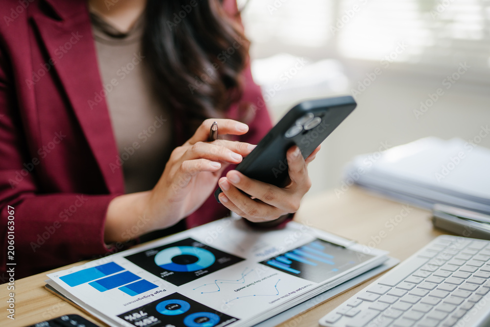 Fototapeta premium Professional woman holding a smartphone and pen, analyzing business charts and data on documents and a digital tablet on a desk
