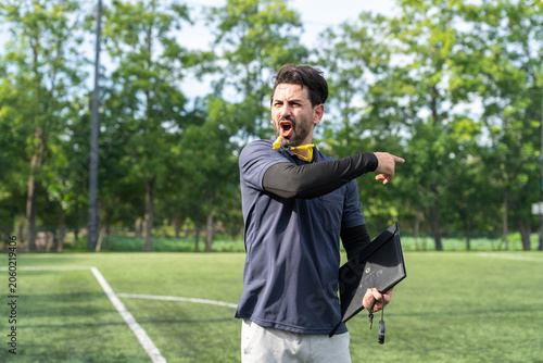 Male sports trainer holding clipboard and whistle yelling instructions inside artificial turf arena, active athletic manager practicing soccer team strategy