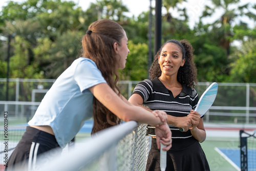 Two young women talking and smiling during pickleball game training session outdoor. Diverse female athletes holding paddle racket near net at court sport lifestyle concept