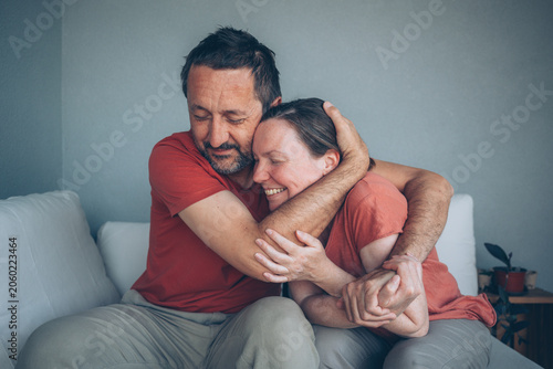 Fotografija A middle-aged couple sits on a couch, embracing with warm smiles and closed eyes, expressing affection, closeness and emotional support in a relaxed, intimate home setting