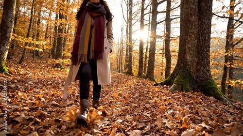 A woman in a cream coat and scarf walks on a leaf-strewn path through a sun-dappled autumn forest, enjoying the season.