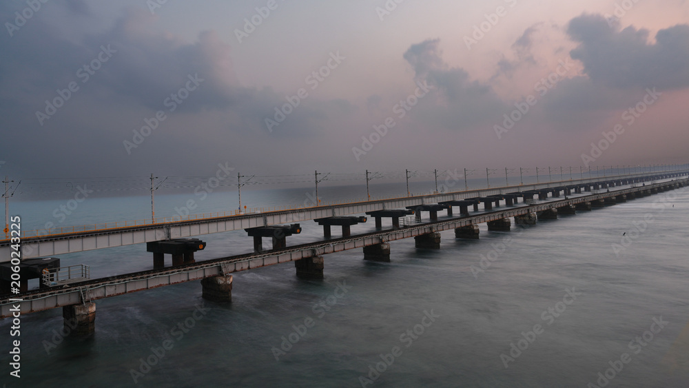 Obraz premium A scenic view of Pamban railway bridge over the sea near Rameswaram in Tamil Nadu, India, captured during soft evening light with cloudy sky and calm ocean water.