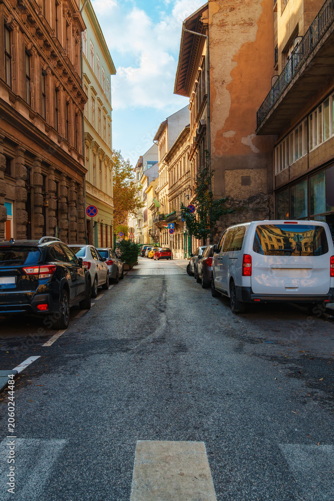 Fototapeta premium Urban street scene with parked cars along a narrow road between historic buildings in Budapest Hungary during daytime with sunlight hitting architecture