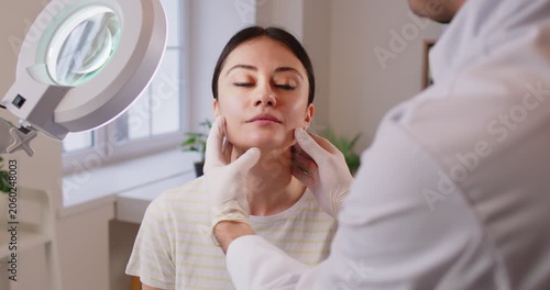 Professional cosmetician examining face skin of young girl in clinic of esthetic cosmetology under special lamp. Plastic surgeon touching woman's face inspecting skin before surgery. 4k video.
