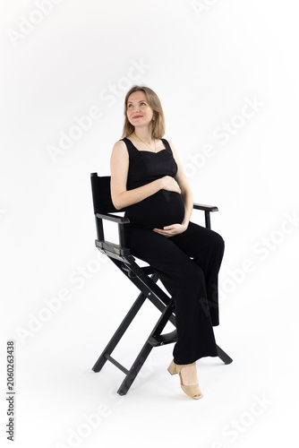 Elegant pregnant woman in black jumpsuit, smiling, sits on a black chair and touch her belly on white background.