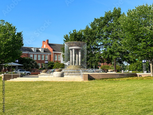 A modern fountain anchors a landscaped plaza at Downtown Cary Park, framed by mature trees, open green space, and historic downtown architecture.