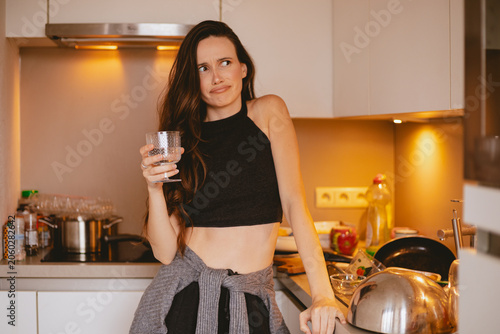 Obraz na płótnie Disgusted and skeptical young woman holding a glass of water, looking away with a repulsed face expression in a messy kitchen with dirty unwashed dishes and pots on the counter
