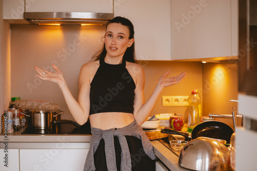 Φωτογραφία Skeptical and unsure young woman shrugging hands while looking at camera with hesitant face expression, standing in messy kitchen surrounded by piles of dirty unwashed dishes and cooking pots