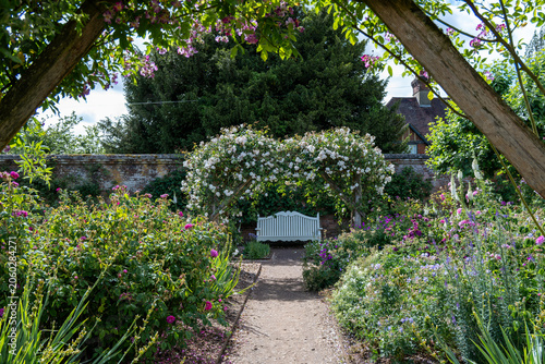 looking down a pathway through a summer garden arbour covered in roses  to a  bench