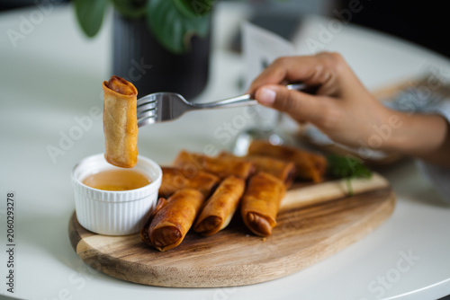 Tableau sur toile Hand of woman using fork for eating fried spring rolls on a wooden kitchen board on white table