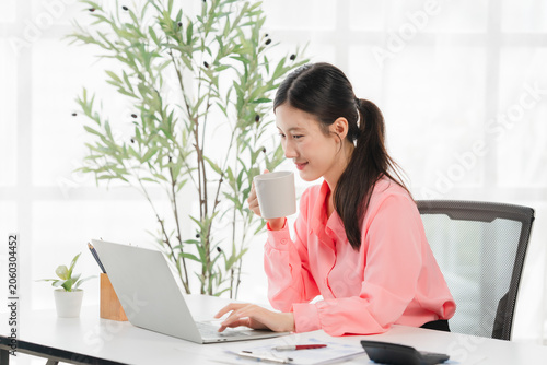 Focused Professional at Work: A young Asian woman in a pink blouse smiles gently as she sips from a white mug while typing on her laptop at a desk.