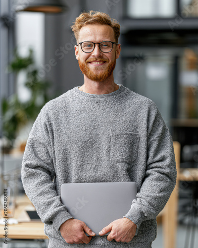 Man with glasses and beard smiles while holding a laptop in a modern office environment.