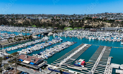 The California, Dana Point Harbor,  with Bots and Yachts in their slips as people interact with the pacific ocean and sea cliffs in the background and sailboats