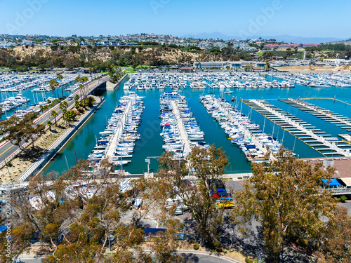 The California, Dana Point Harbor,  with Bots and Yachts in their slips as people interact with the pacific ocean and sea cliffs in the background and sailboats