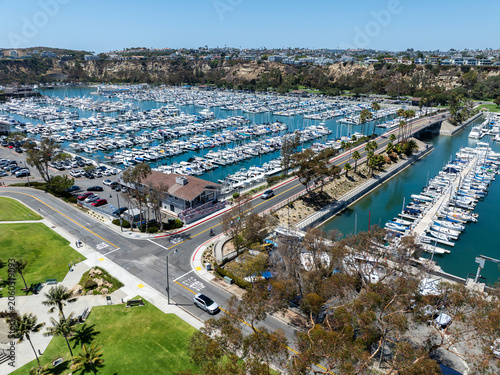 The California, Dana Point Harbor,  with Bots and Yachts in their slips as people interact with the pacific ocean and sea cliffs in the background and sailboats