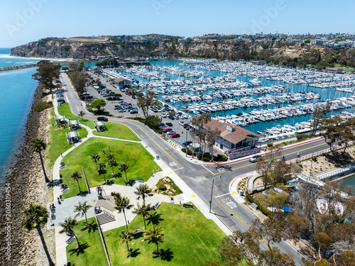 The California, Dana Point Harbor,  with Bots and Yachts in their slips as people interact with the pacific ocean and sea cliffs in the background and sailboats with the Richard Henry Dana Monument 