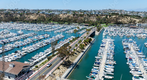 The California, Dana Point Harbor,  with Bots and Yachts in their slips as people interact with the pacific ocean and sea cliffs in the background and sailboats