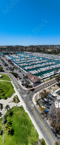 The California, Dana Point Harbor,  with Bots and Yachts in their slips as people interact with the pacific ocean and sea cliffs in the background and sailboats
