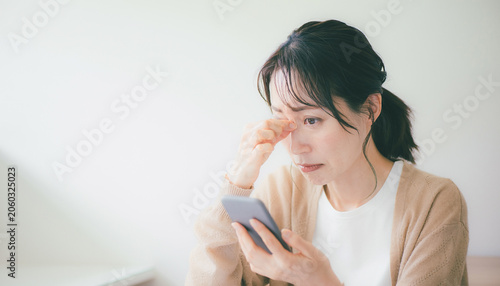 Stressed Japanese woman looking at smartphone with tired eyes スマホを見て目が疲れ頭を悩ませる日本の女性