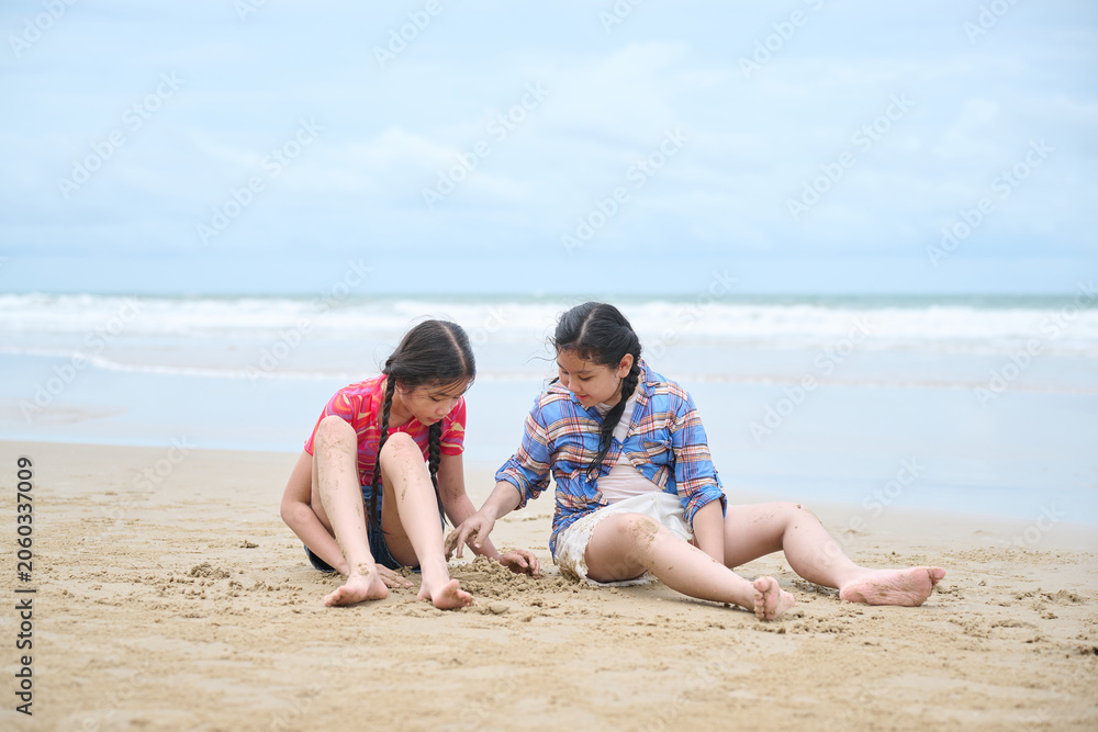 Fototapeta premium Girls sitting on a sandy beach, playing with the sand. They are both looking down at their hands as they build with the sand. The water is in the background.