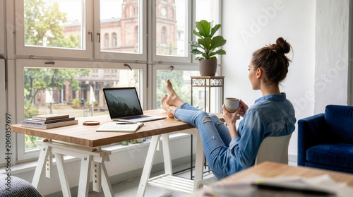 Young woman relaxing with bare feet on a wooden desk, holding coffee and looking out the window while remote working on a laptop in a bright home office.