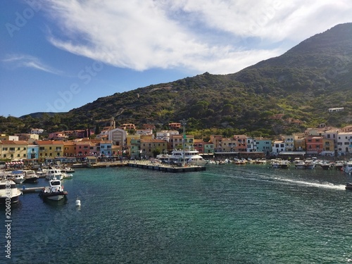 Puerto marítimo de Isola del Giglio con casas de colores y barcos amarrados bajo un cielo azul, Toscana, Italia.