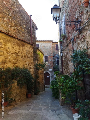 Callejón medieval empedrado con casas de piedra, plantas en macetas y farola clásica en Montemerano, Toscana, Italia.
