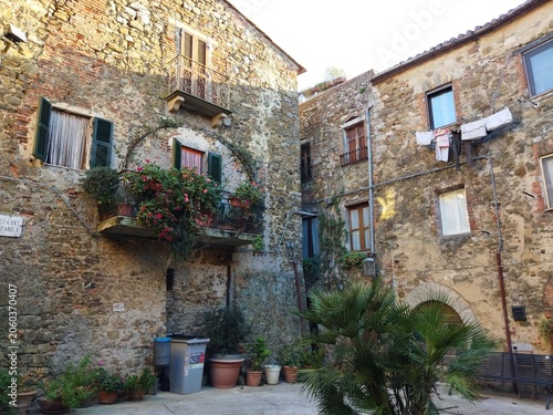 Plaza medieval con fachadas de piedra históricas, balcón decorado con flores y ropa tendida en Montemerano, Toscana, Italia.