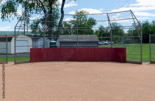 Bild auf Leinwand Empty baseball or softball field with a protective chain-link backstop and red padding, featuring a dirt infield, green outfield, sheds, and blue sky with clouds on a sunny day