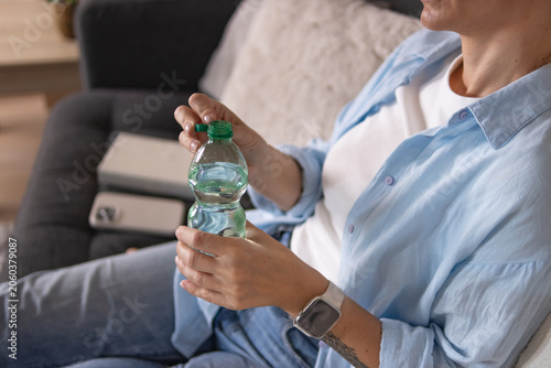 Canvastavla Woman sitting on sofa at home drinking water from bottle, relaxing and rehydrating in comfortable living room after a warm day, enjoying calm indoor lifestyle moment