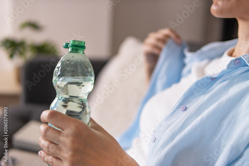 Obraz na plátně Woman sitting on sofa at home drinking water from bottle, relaxing and rehydrating in comfortable living room after a warm day, enjoying calm indoor lifestyle moment