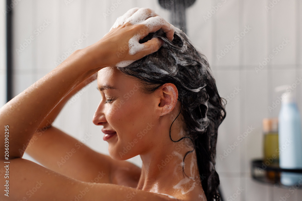 Fototapeta premium Side view of lady applying shampoo and washing hair taking shower in modern bathroom, haircare routine using cosmetics
