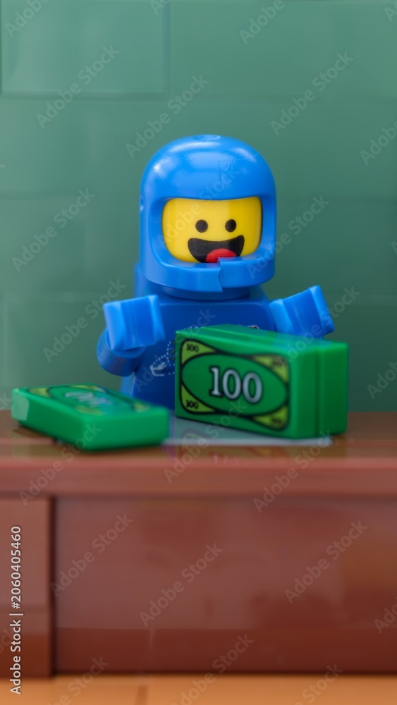 Naklejka premium Tambov, Russian Federation - May 26, 2025 Close-up shot of a blue Lego astronaut minifigure counts green money at a brown wooden desk.