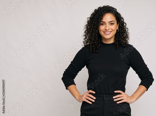 Confident young woman with curly hair smiling. Female in black turtleneck standing with hands on hips. Professional portrait on grey background with copy space
