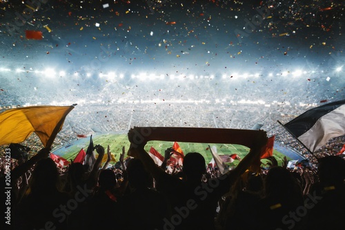 Soccer crowd cheering in stadium at championship final match. Supporters holding flags and scarf during sports tournament victory. Emotional fan atmosphere with flying confetti at night