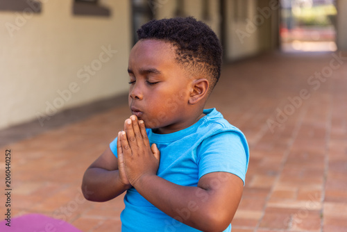 African American boy kneeling in tile hall praying in bright-blue-tee near purple mat, copy space
