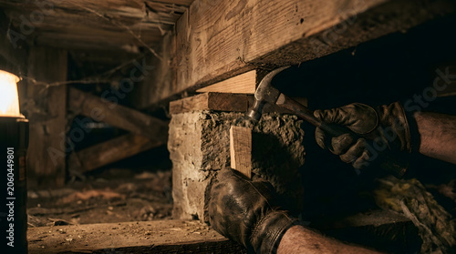 A construction worker uses a hammer to drive a wooden shim into the stone foundation of a house in a dark, dimly lit crawlspace, illustrating home repair, renovation, and structural maintenance work.