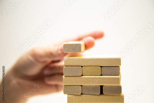 Close-up of the top tower made of wooden bars, a game of jengo, standing on a white background, hand on top placing a wooden block. The concept of board games, business, construction. High quality
