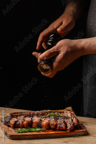 Chef seasoning sliced porterhouse steak with pepper on wooden serving board