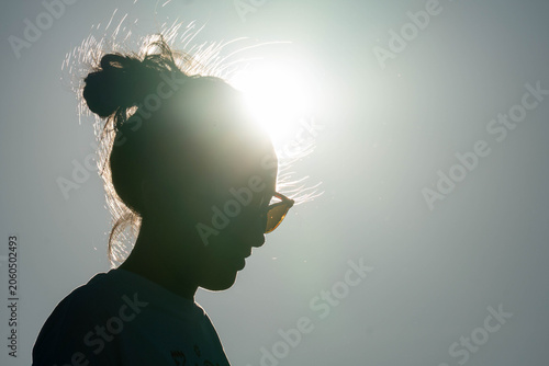 Silhouette of a woman in sunglasses with a messy bun, backlit by bright sun against a clear sky, creating a dramatic summer profile.